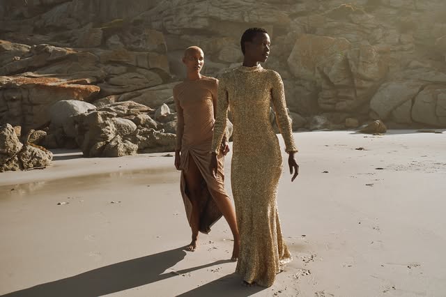 Two fashion models in long dresses on a sandy beach beside sculpted rocks